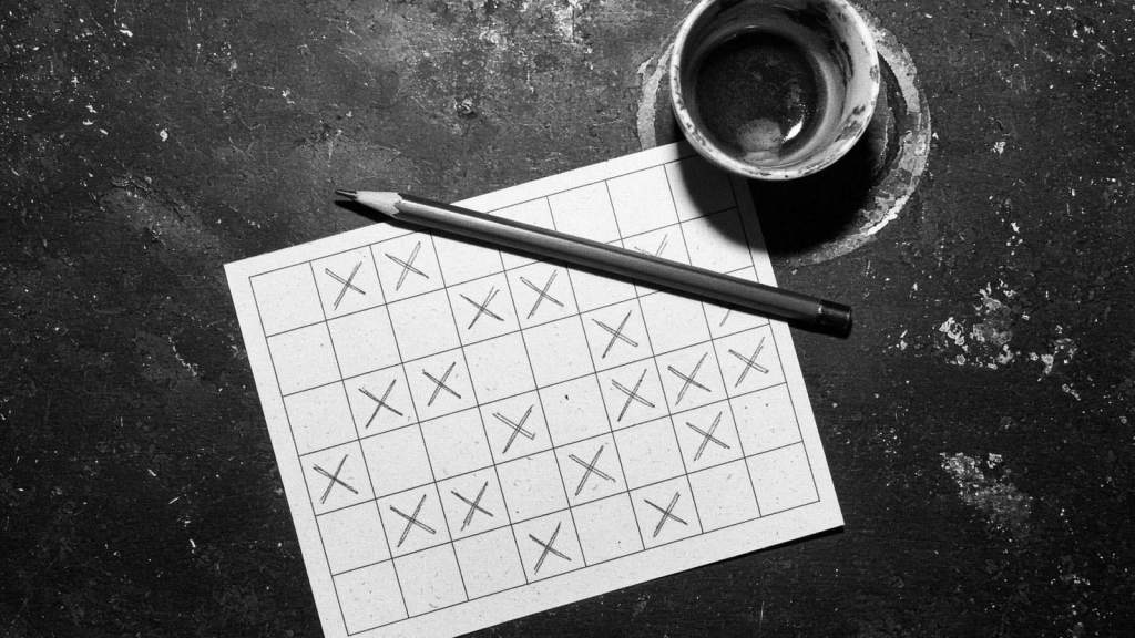 A printed grid card filled in with pencilled crosses, lying on a scuffed dark surface with a pencil across it and an empty coffee cup alongside.