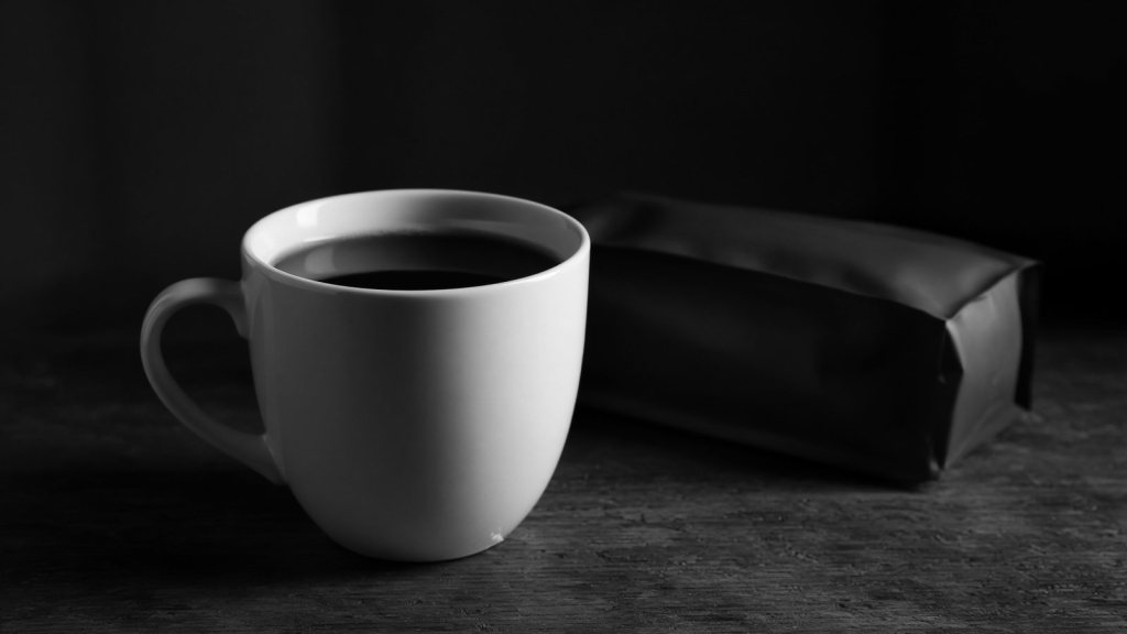 A plain white cup of black coffee on a dark wooden surface, with an unlabelled coffee bag lying face down behind it.